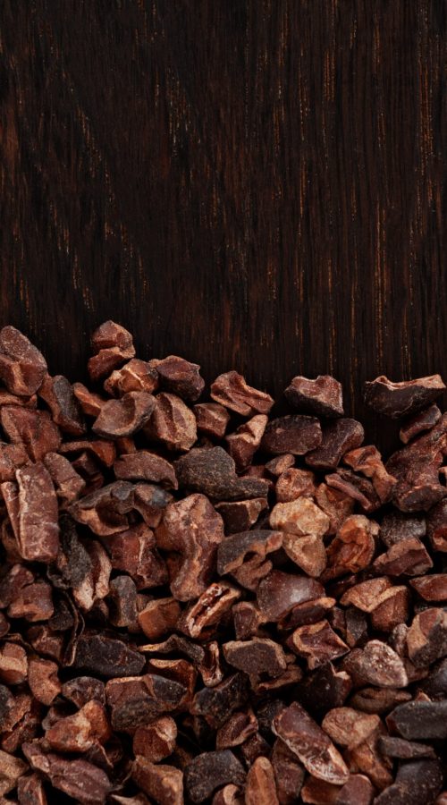 Cocoa nibs on brown wooden board, close-up. Design element, copy space. Crunchy pieces of peeled, crushed and lightly roasted cocoa beans with a pleasant chocolate bitterness.